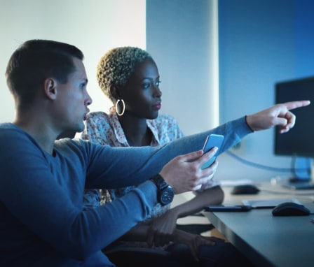 Two people looking at code on a desktop and holding a mobile phone