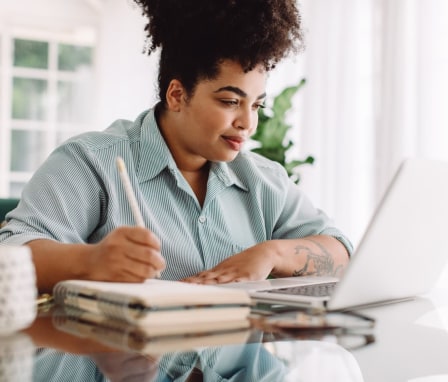 Woman taking notes in notebook while reading on laptop at home